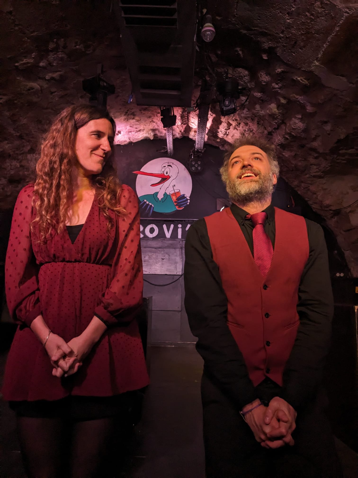 Une femme et un homme se tiennent dans une cave sombre ornée d’un décor de Noël, avec un dessin d’un Flamand rose portant un verre présenté en arrière-plan.