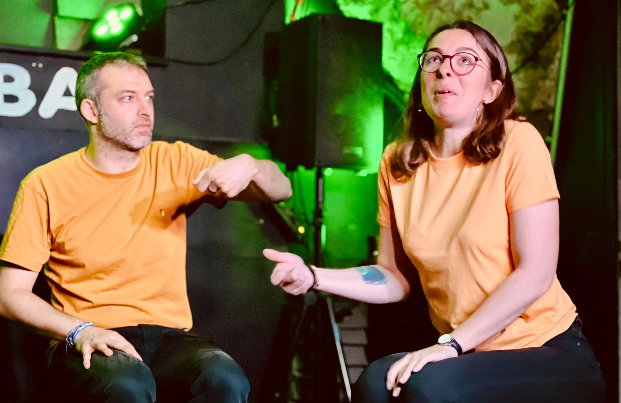 Deux personnes en conversation, une homme et une femme, portant des T-shirts orange, assises dans un environnement avec des lumières vertes.