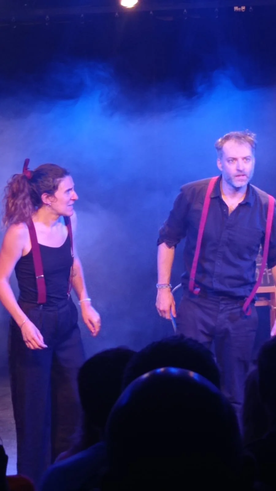 Deux acteurs sur scène durant une performance théâtrale, l'homme porte des bretelles rouges et une chemise sombre, la femme porte un haut noir et a les cheveux attachés avec un ruban rouge.