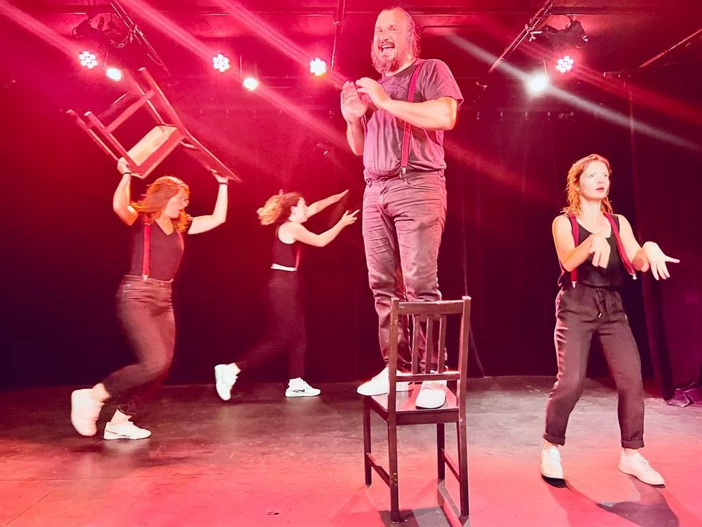 Scène de théâtre avec cinq acteurs : une femme avec un fauteuil, deux femmes qui dansent, une femme avec un bras levé, un homme debout sur une chaise, tous sous un éclairage rouge.