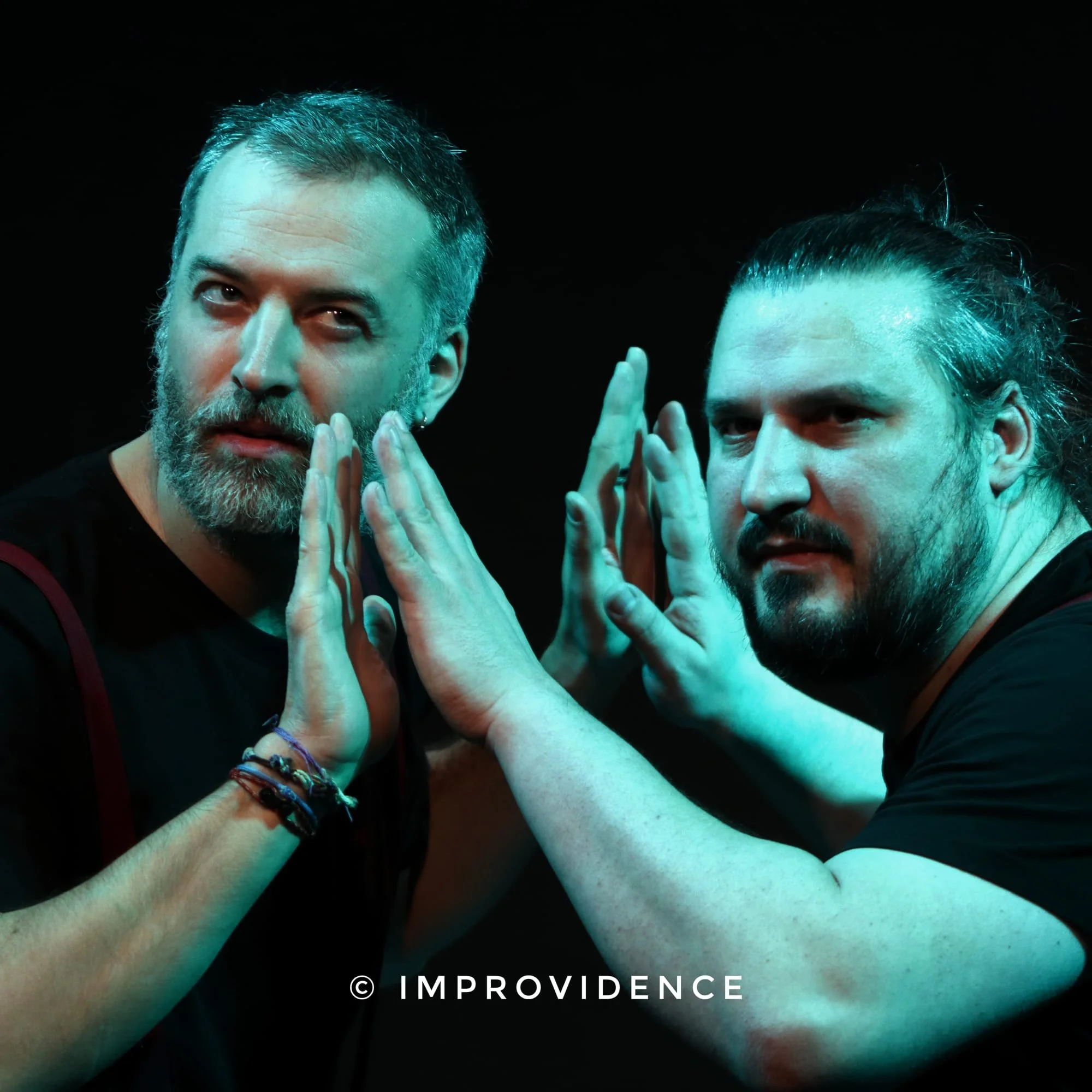 Deux hommes avec des barbes et des cheveux longs, en train de faire un geste avec leurs mains, dans une scène sombre avec un éclairage bleu.