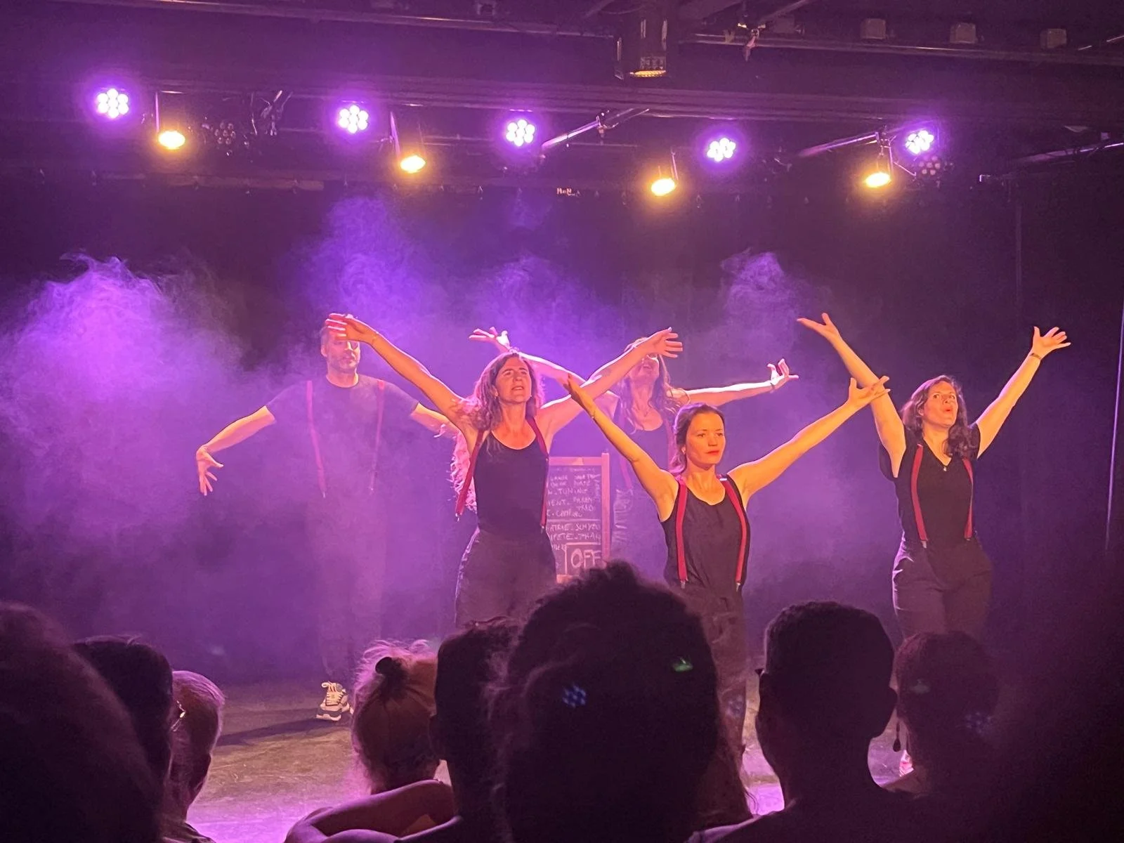 Quatre danseuses en costume noir avec des bretelles rouges effectuent une performance sur scène sous des éclairages violets et jaunes, entourées de fumée, avec un public regardant.