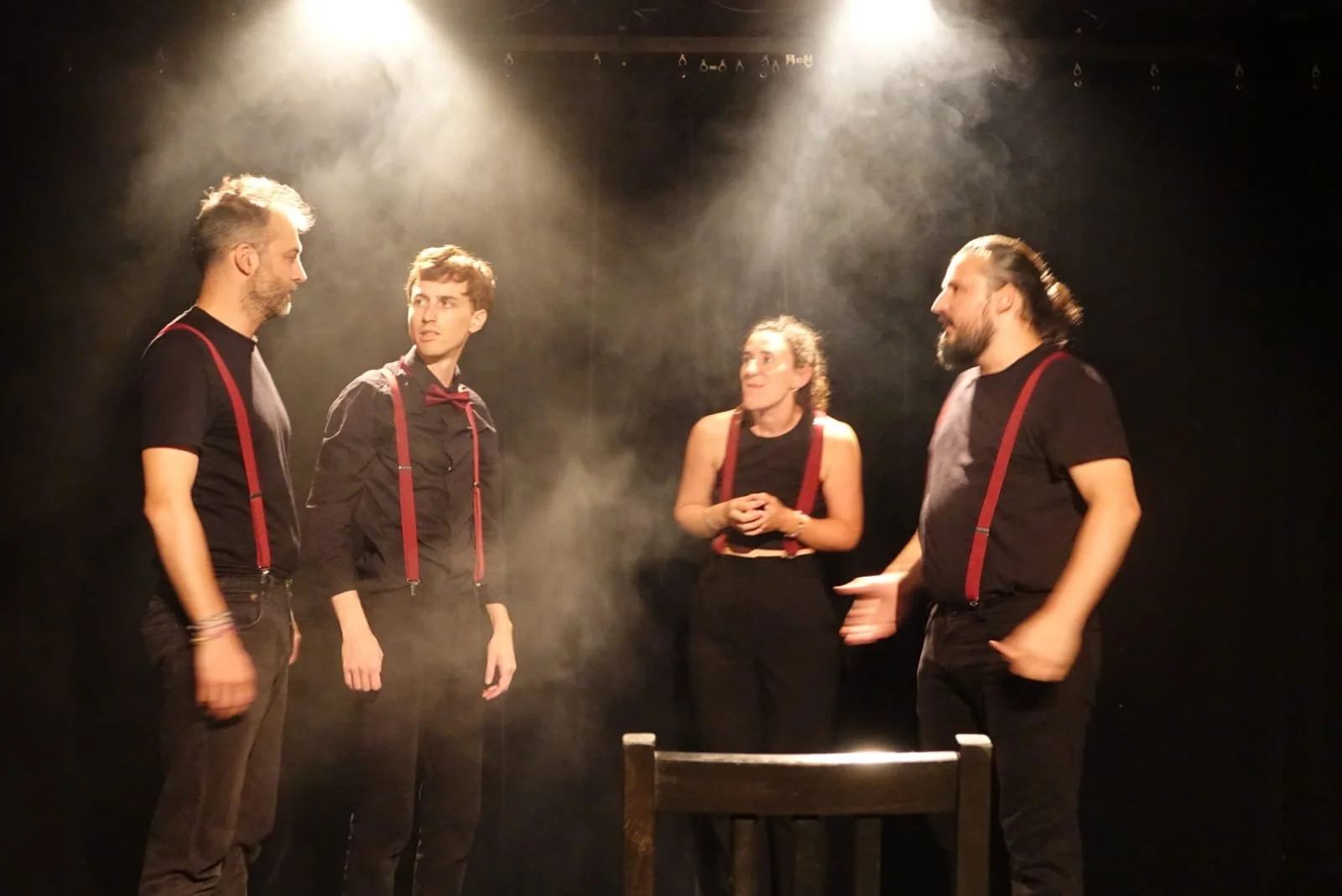 Quatre personnes sur scène, discutant, avec un éclairage doux et de la fumée, portant des vêtements noirs avec des bretelles rouges.