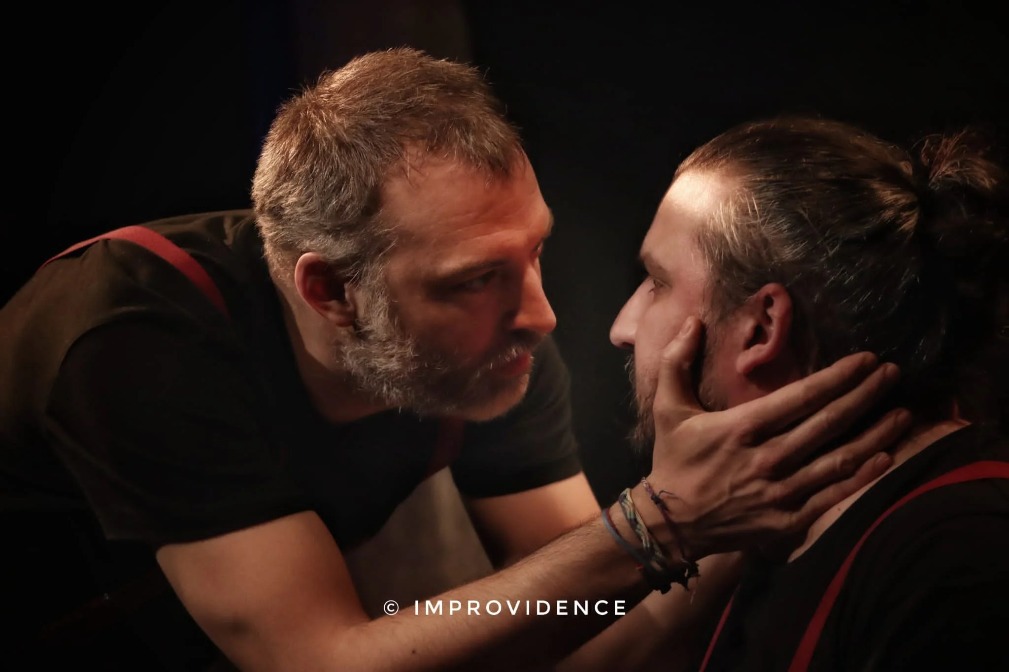 Deux hommes regardent intensément, l'un tenant le visage de l'autre avec les mains. L'ambiance est sombre et dramatique.