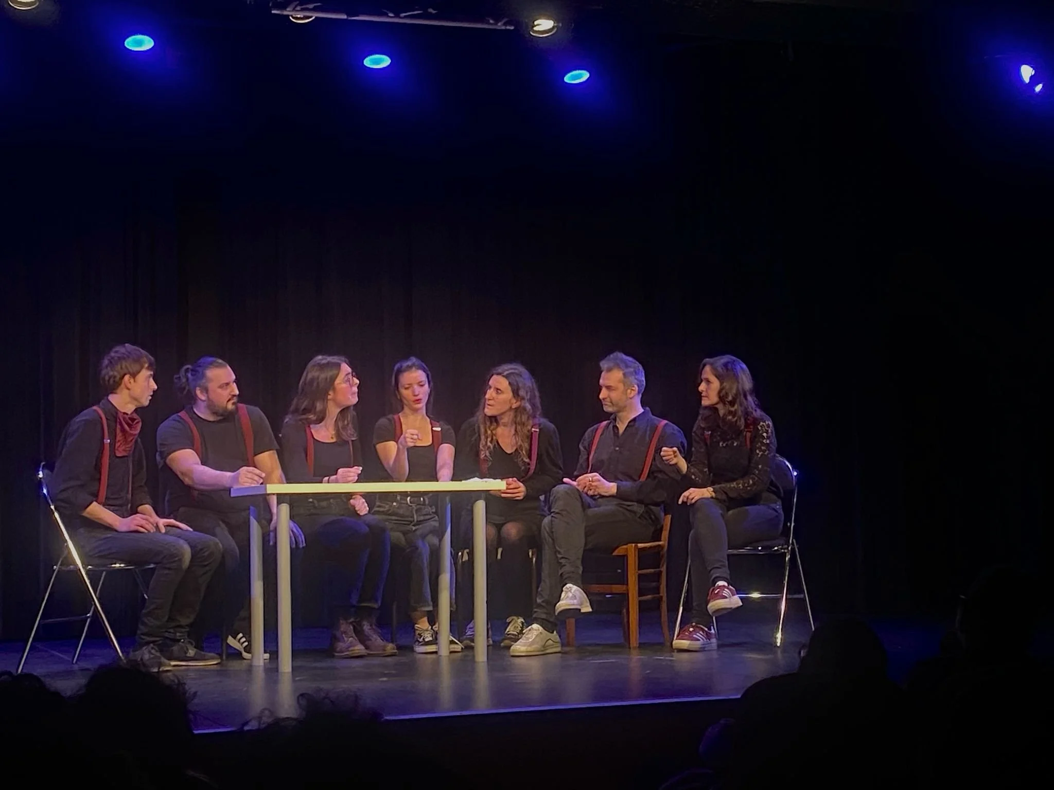 Groupe de sept acteurs sur scène lors d'une pièce de théâtre, assis autour d'une table.