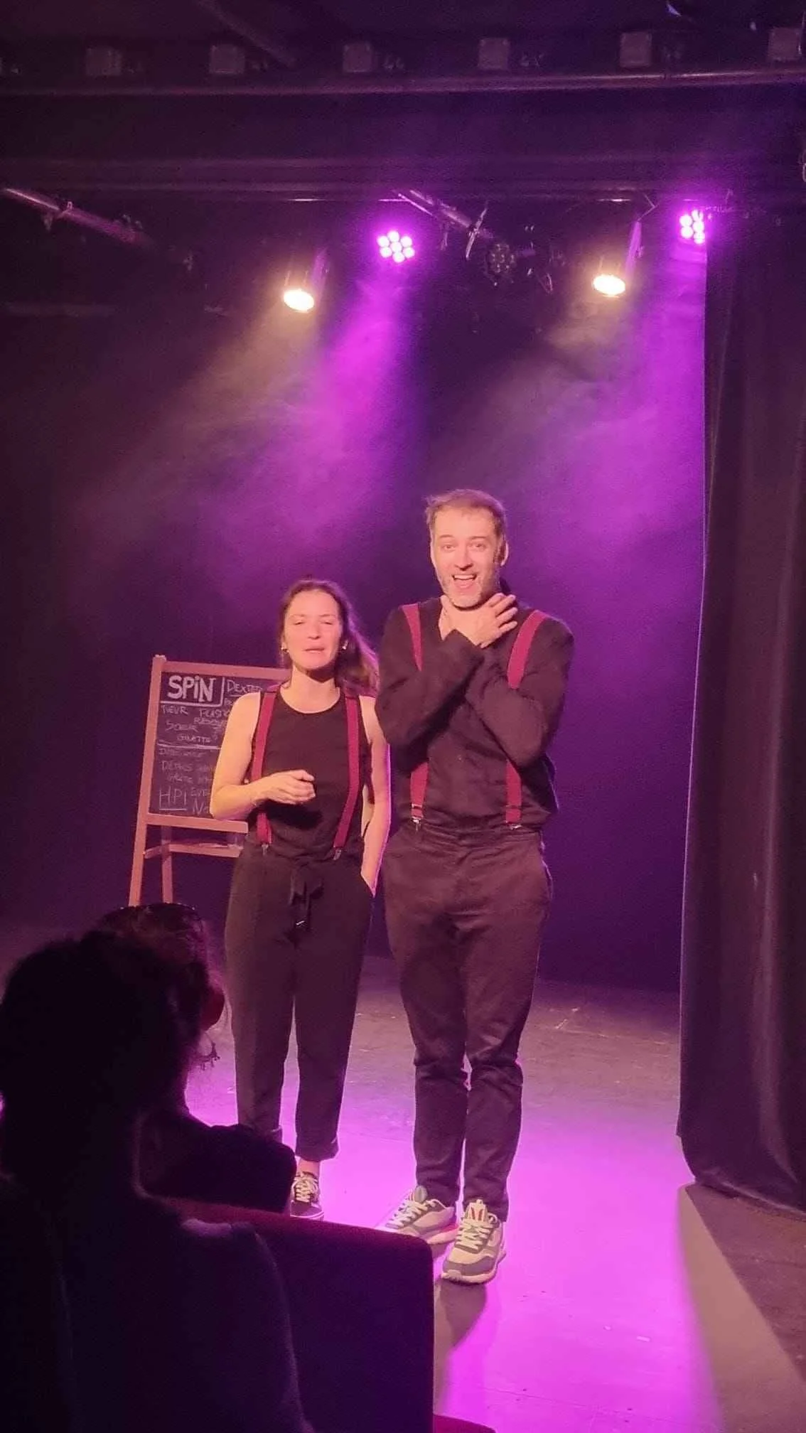 Deux comédiens sur scène, un homme et une femme, sous des lumières pourpre, avec un panneau noir en arrière-plan.