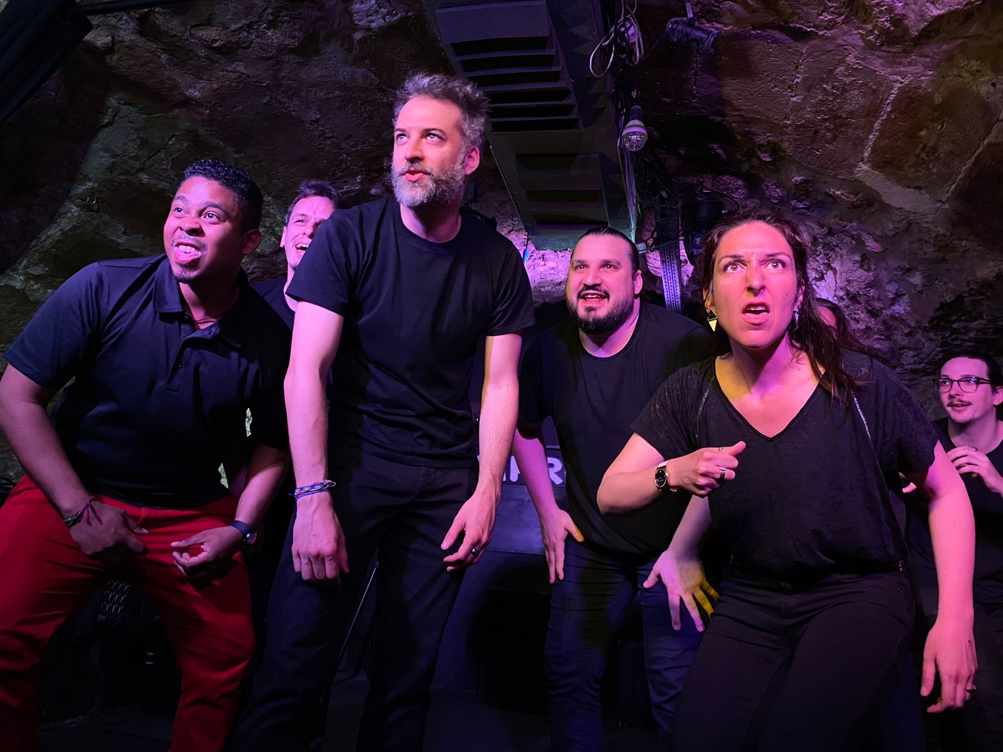 Un groupe de personnes sur scène, ayant des expressions de surprise ou de confusion, dans une ambiance sombre avec un éclairage violet.