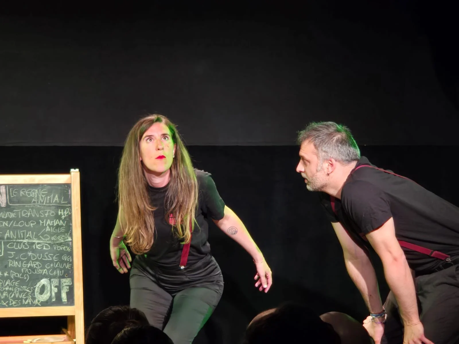 Deux comédiens en costume noir jouent une scène dramatique sur scène noire, avec un tableau noir à gauche.