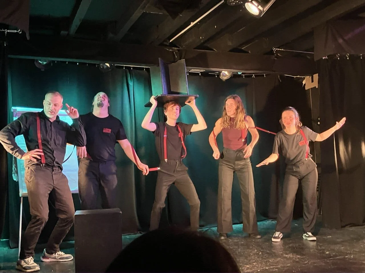 Groupe de cinq personnes sur scène, en train de faire une performance comique ou artistique, avec un fond noir et des éclairages de scène.