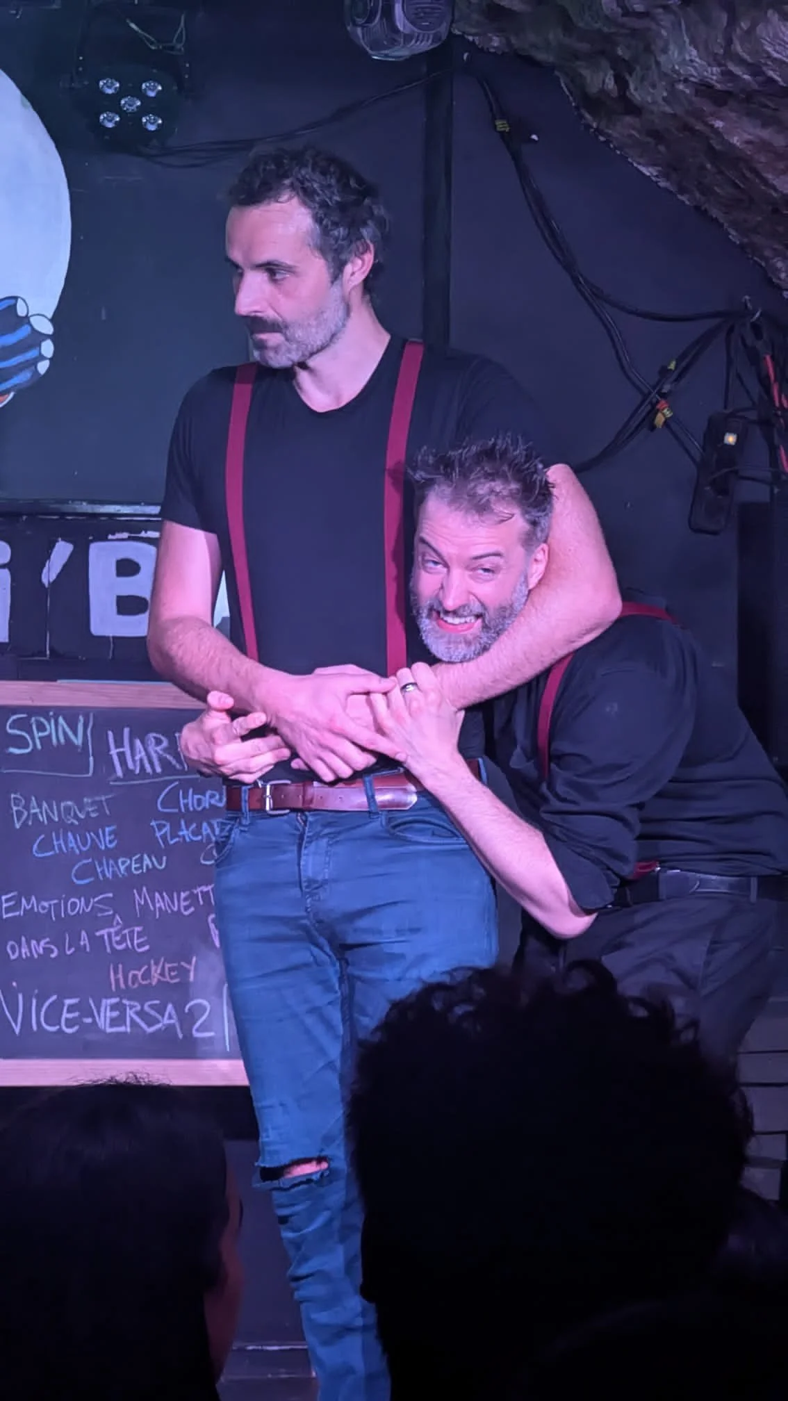 Deux hommes jouent à un jeu ou mimique sur scène, l'un semble en position de domination ou d'humour, tandis que l'autre sourit ou grimace, dans un café ou une salle de spectacle, avec un tableau de menu en arrière-plan.