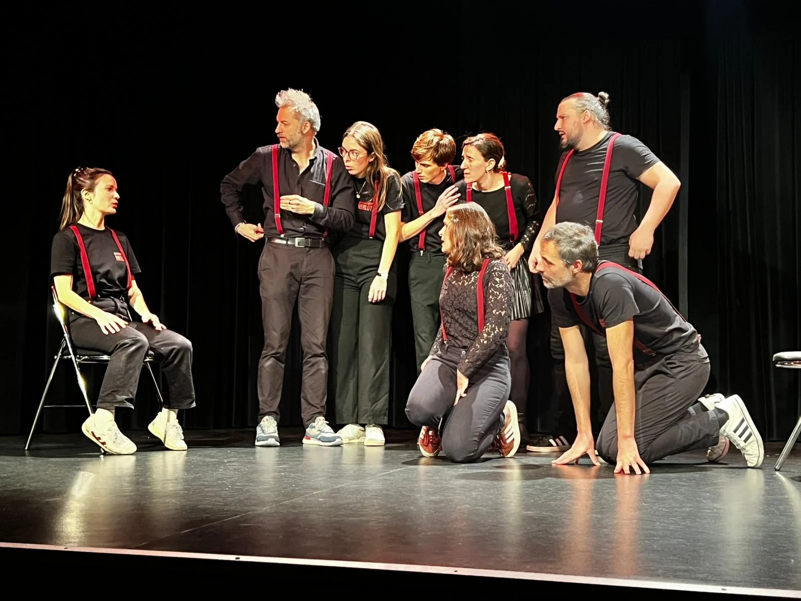 Groupe de huit acteurs en costume noir avec suspendeurs rouges pendant une pièce de théâtre sur scène, certains assis ou à genoux, d'autres debout, discutant et jouant leurs rôles.