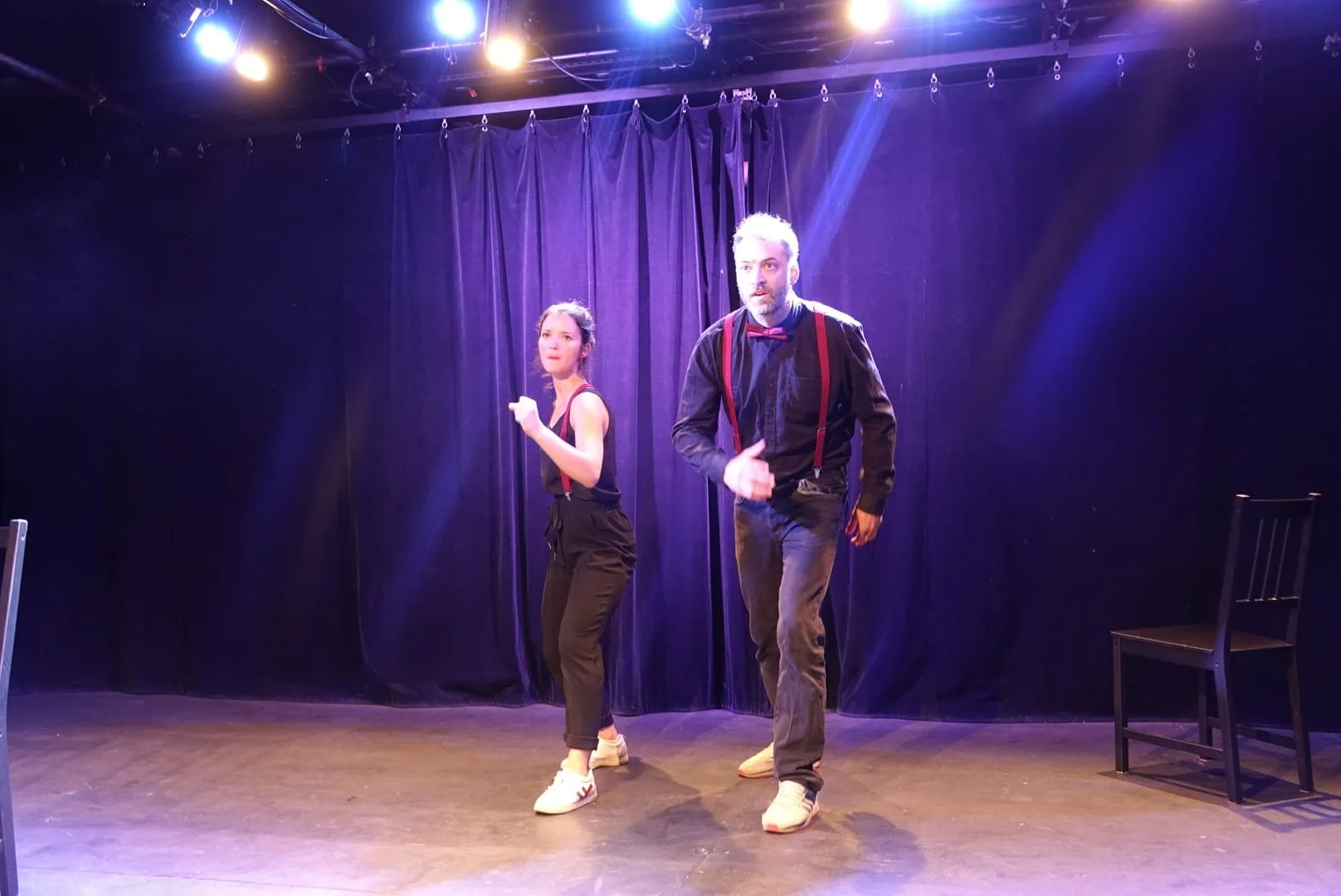 Deux acteurs sur scène, une femme et un homme, en train de jouer une scène de théâtre avec un fond de rideau violet et des éclairages. L'homme porte une chemise noire avec des bretelles rouges et un nœud papillon rouge, et la femme porte un débardeur