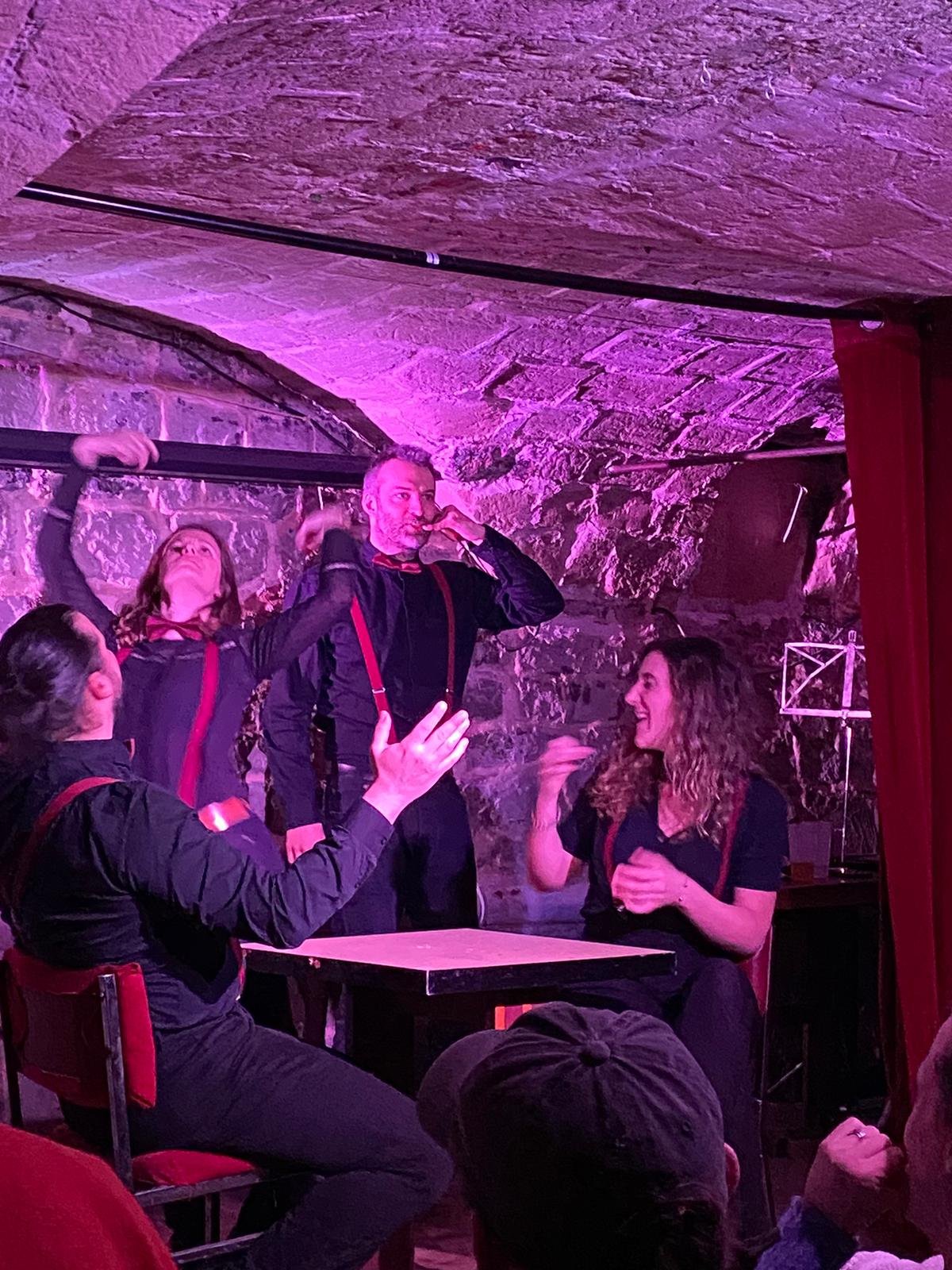 Quatre personnes en costume de scène noir avec des suspentes rouges, performants dans un spectacle de théâtre dans un lieu souterrain avec des murs en pierre, éclairé par des lumières roses ou violettes.