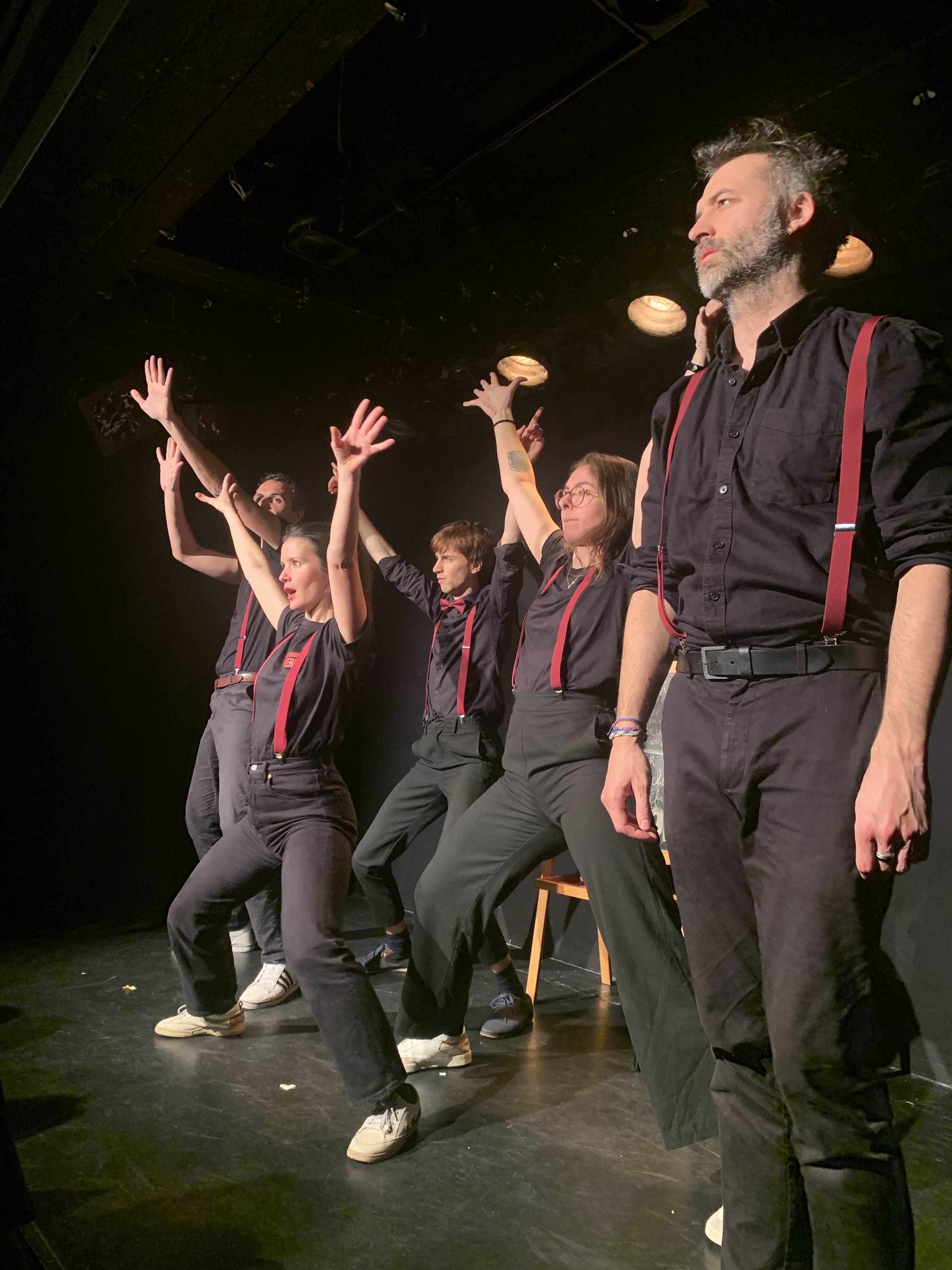 Groupe de cinq personnes en costume noir avec bretelles rouges sur scène, posant ou en mouvement, sous un éclairage de théâtre.