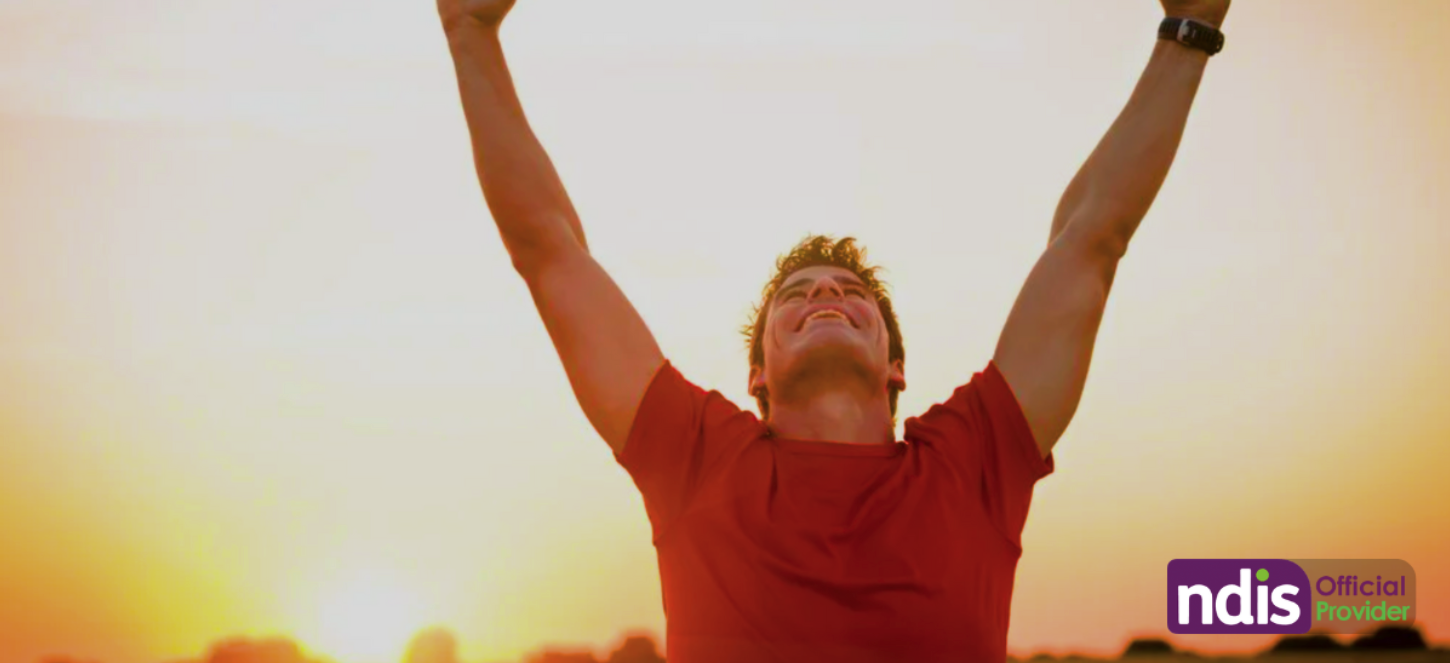 A person wearing a red shirt and black watch smiling with arms raised upward outdoors at sunset.