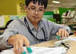 A young man sitting at a desk in an office, looking at papers and using a calculator.