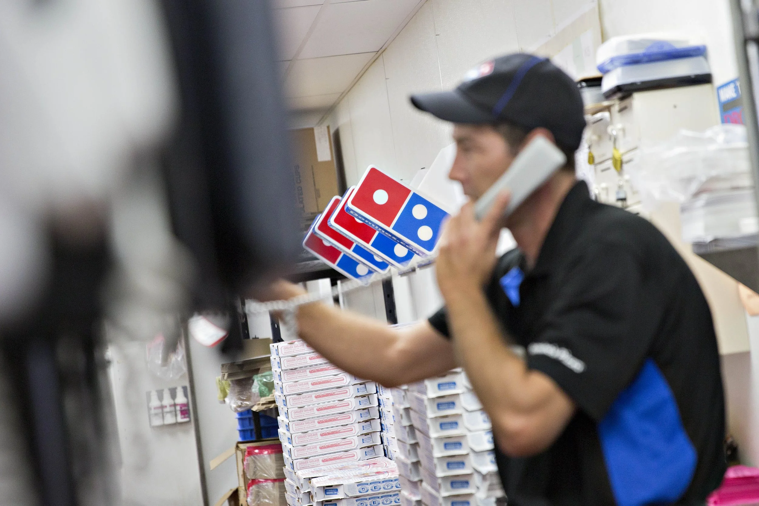 A person in a Domino's Pizza uniform talking on a landline phone inside a store, with pizza boxes and supplies in the background.