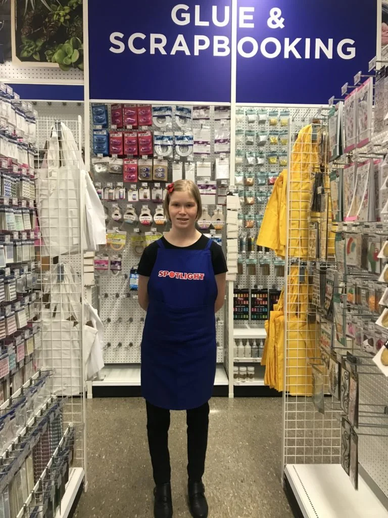 A young girl wearing a blue apron standing in a store aisle labeled 'Glue & Scrapbooking'. She is smiling with hands behind her back, surrounded by various art supplies, scrapbooking materials, and yellow raincoats.