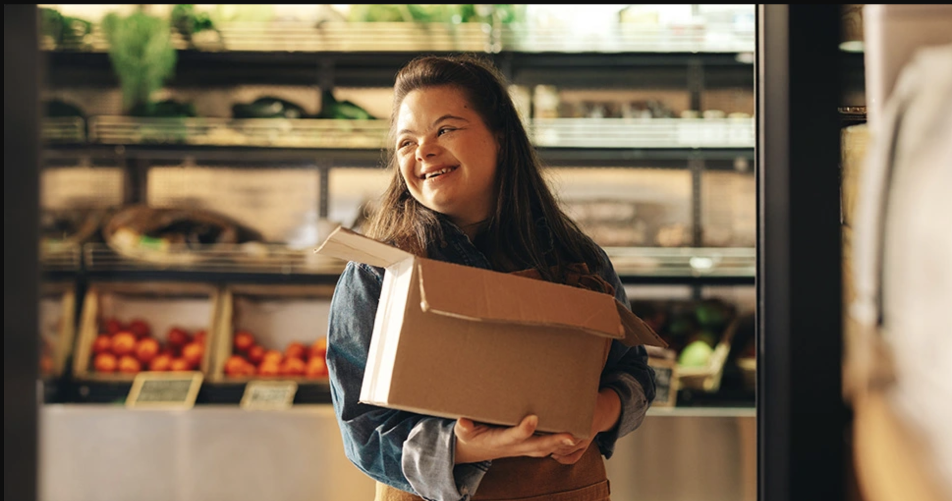 A young woman with long dark hair smiling while holding a cardboard box of groceries inside a store near fresh produce shelves.