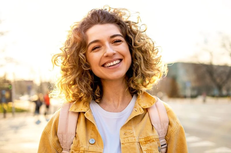 A young woman with curly hair smiling outdoors during sunset, wearing a yellow jacket and carrying a backpack.
