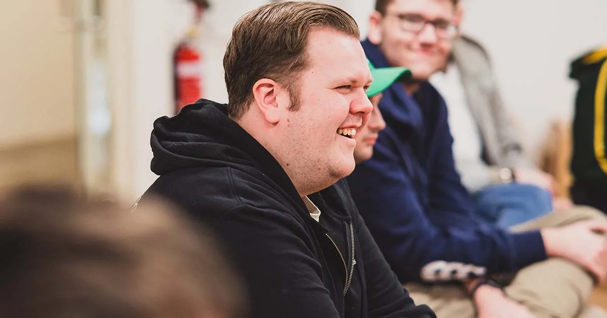 A group of men sitting in a row, smiling and engaging in conversation, in an indoor setting.