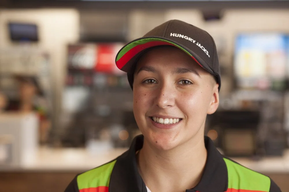 Young man smiling, wearing a black cap with green and red accents and the words 'Hungry Jack's', in a fast-food restaurant.