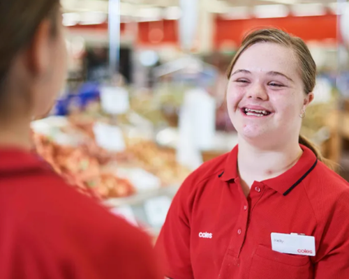 Smiling young girl working at Coles grocery store counter, talking to a customer.