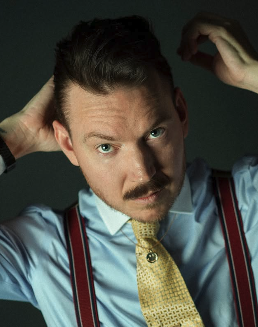 A man with dark hair and a mustache wearing a light blue dress shirt, yellow patterned tie, and red suspenders, looking directly at the camera with his hands behind his head.