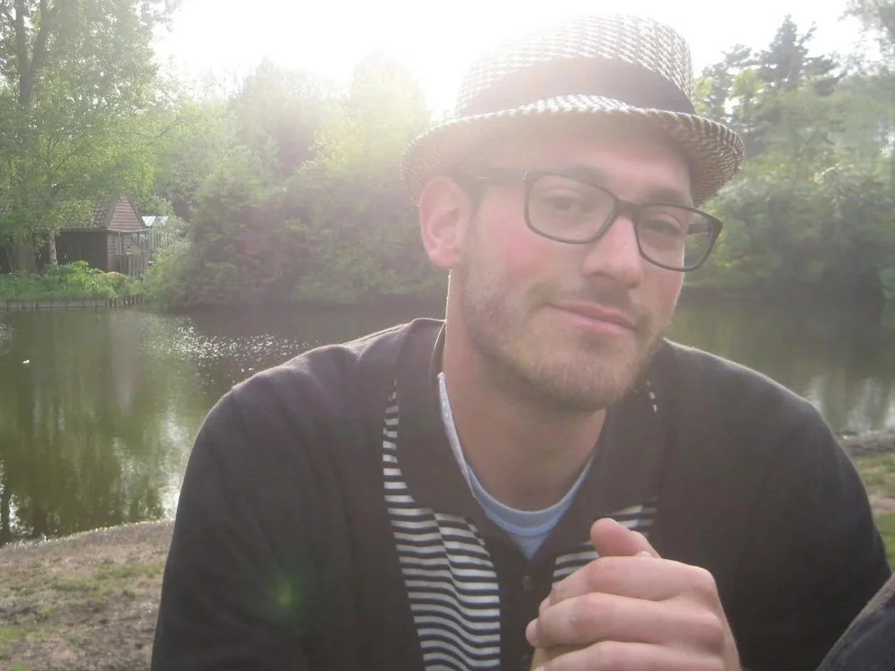 Yehudah Sunshine, wearing glasses, a fedora hat, and a striped shirt, is sitting outdoors near a body of water with lush green trees and a small wooden house in the background.