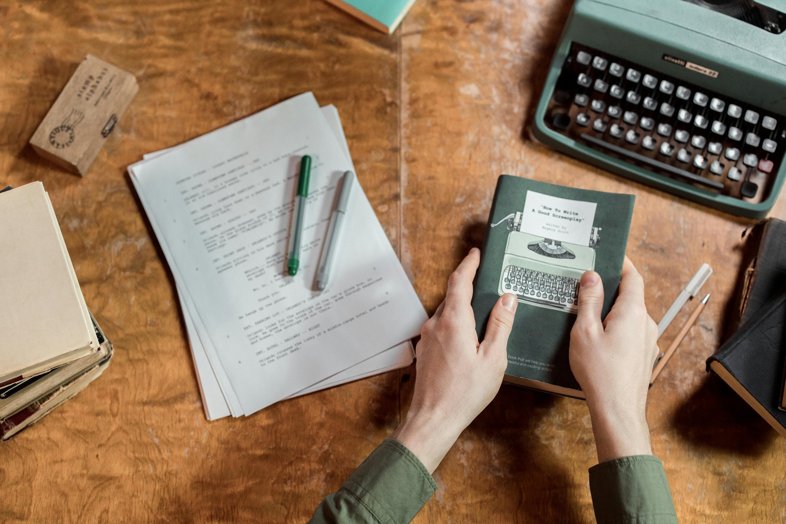 Person holding a book titled "How to Write a Good Screenplay" with a typewriter illustration on the cover, surrounded by papers, pens, a typewriter, and other books on a wooden desk.