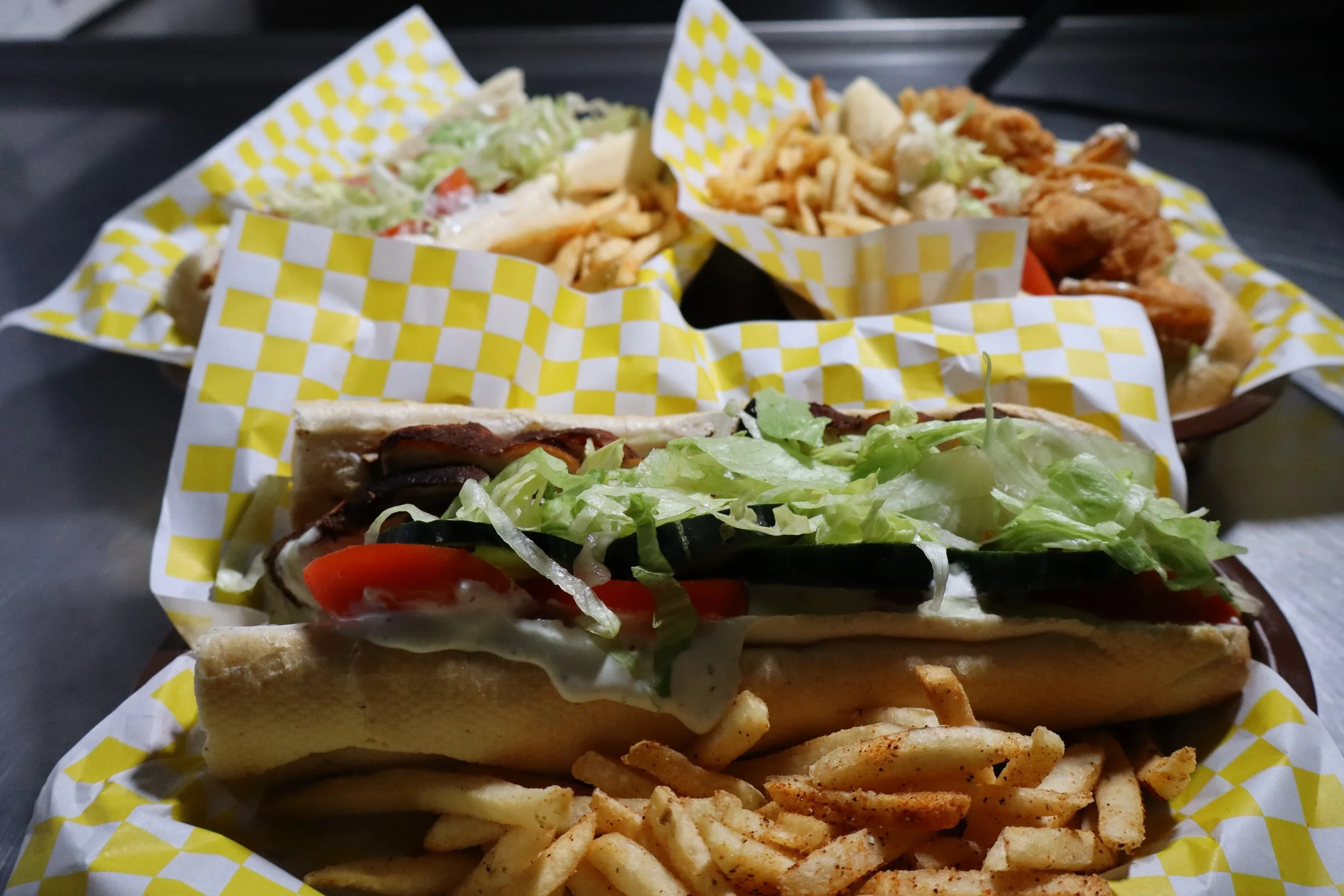 Three sandwiches with lettuce, tomato, and other vegetables, served with French fries and coleslaw on yellow checkered paper.