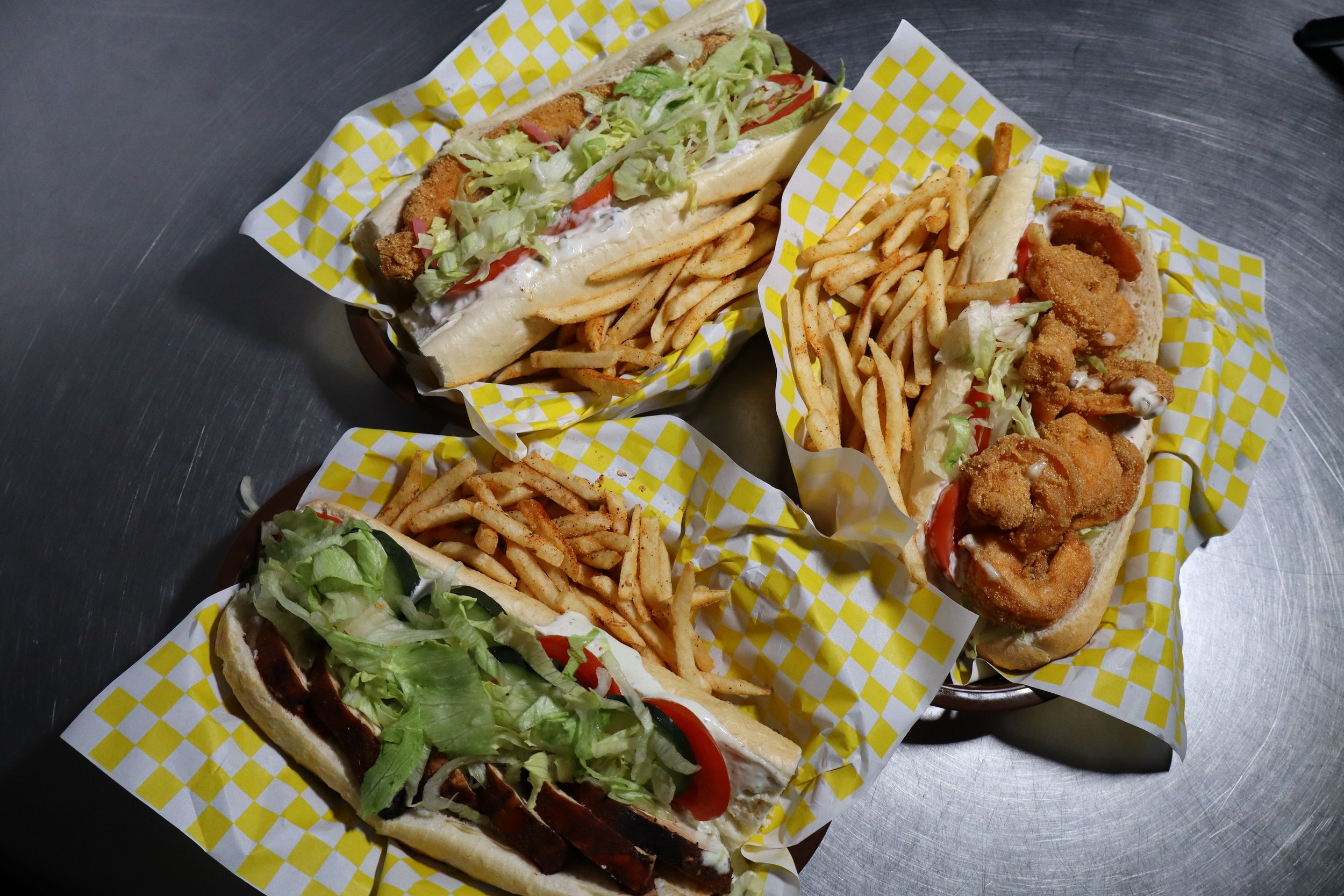 Three baskets with fast food sandwiches and fries on a metal table. The sandwiches include fried fish, fried chicken, and sub with vegetables.