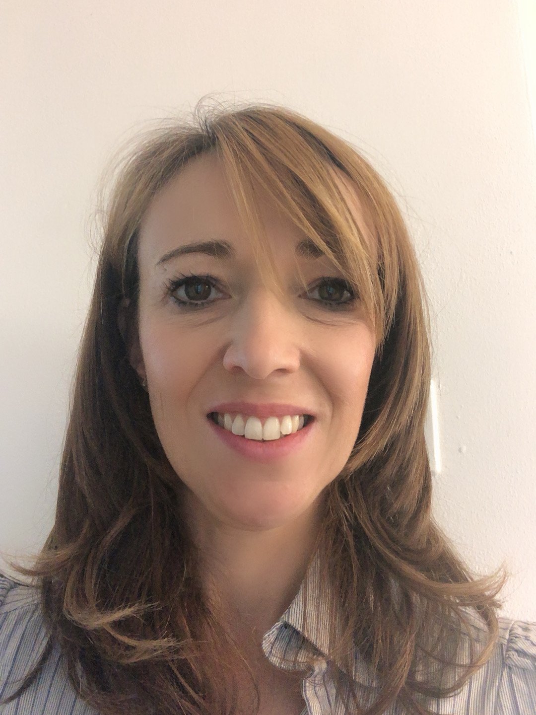 Primer piano di una donna con capelli castani chiari e occhi chiari, che sorride, indossa una camicia a righe, sfondo neutro.