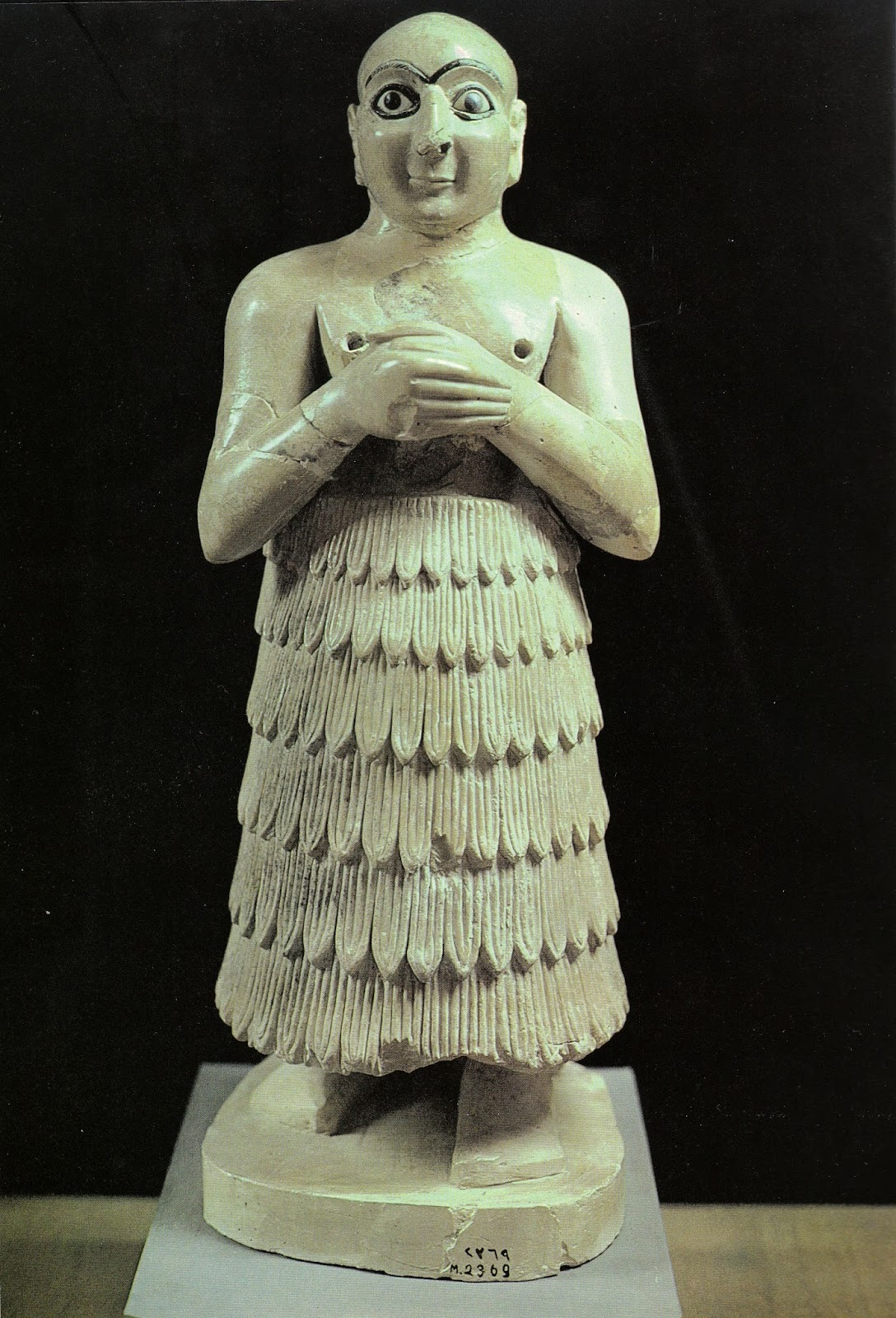 Ancient statue of a standing figure with hands crossed over the chest, wearing a layered skirt made of carved leaves, with a round base, in a museum setting.