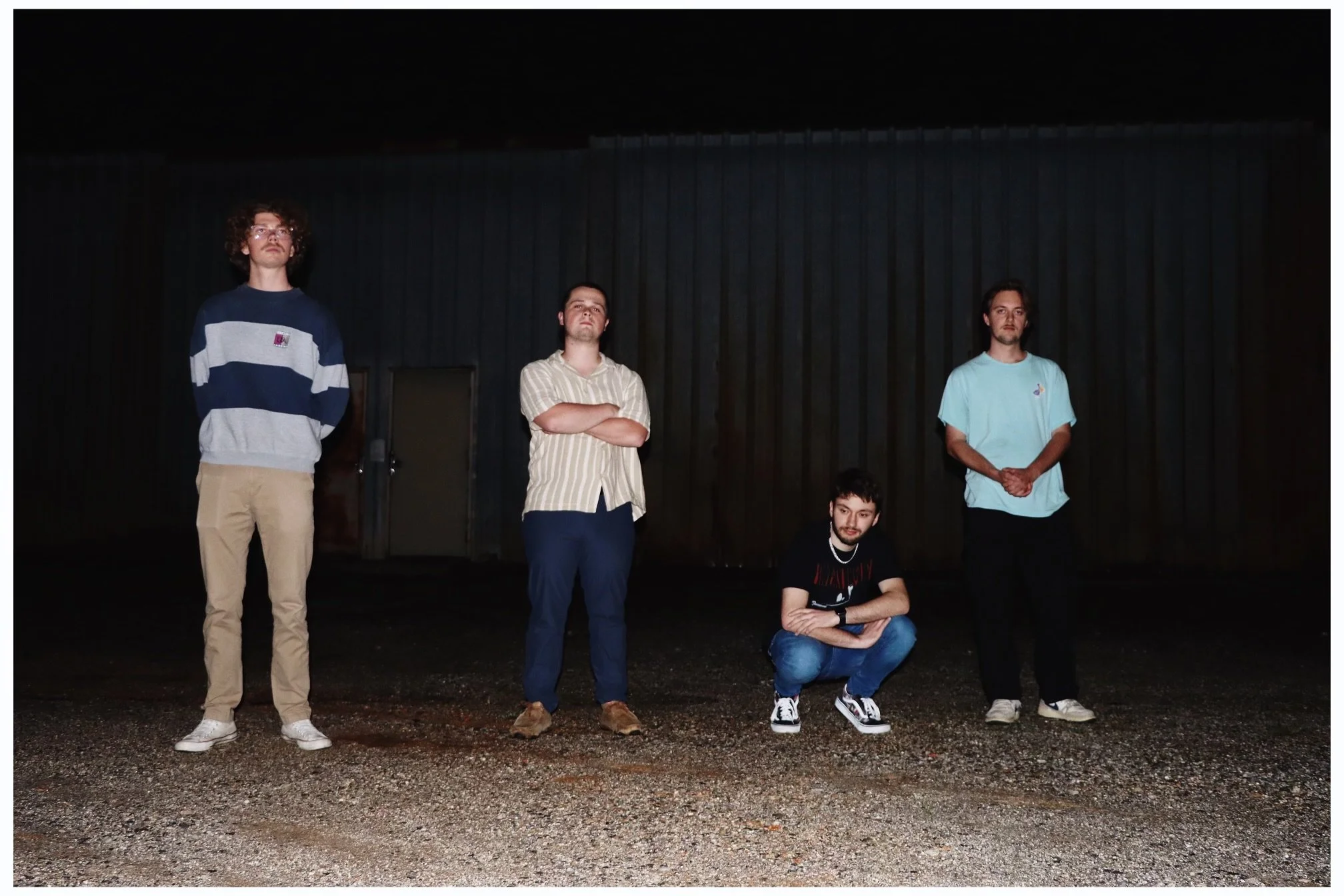 Five young men standing and crouching outdoors at night in front of a dark metal wall, with gravel ground.