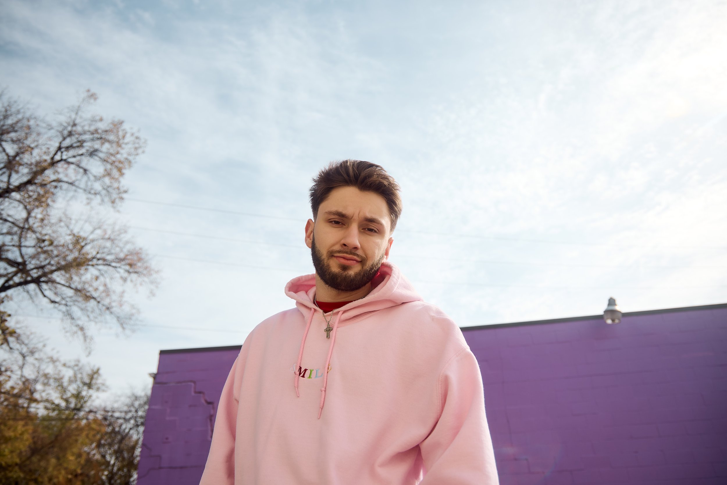 A young man with dark hair and a beard wearing a pink hoodie with the word 'SMILE' embroidered on it, standing outdoors with trees and a purple building behind him under a partly cloudy sky.