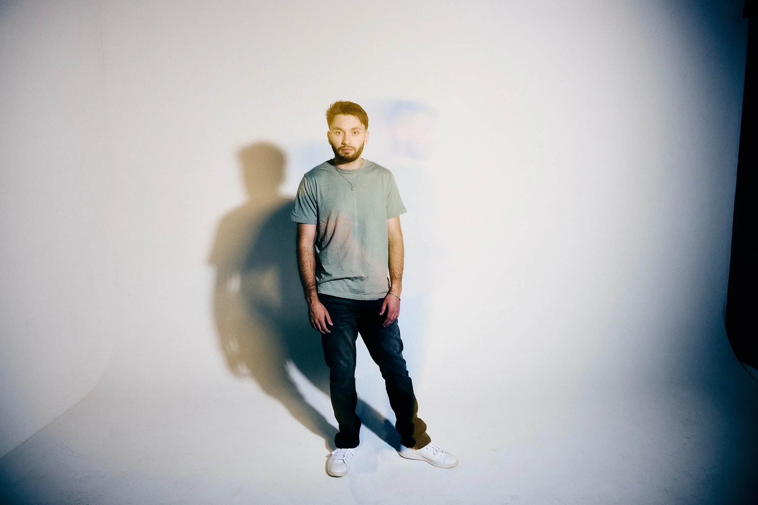 A young man standing in front of a plain white background with a shadow cast behind him. He has short brown hair and a beard, wearing a gray t-shirt, dark jeans, and white sneakers.