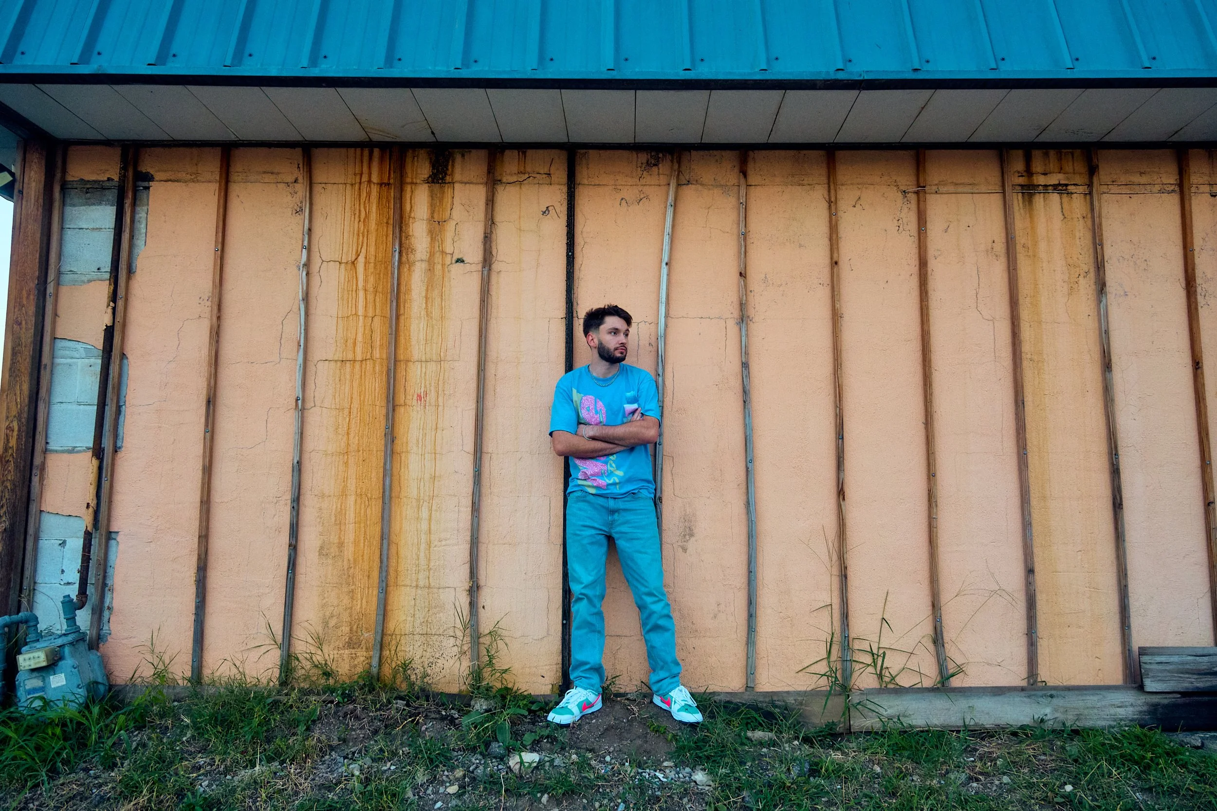 A young man with a beard stands with arms crossed against a partially exposed wall with vertical wooden studs and peeling paint, wearing a bright blue T-shirt, matching blue pants, and white sneakers with pink accents.