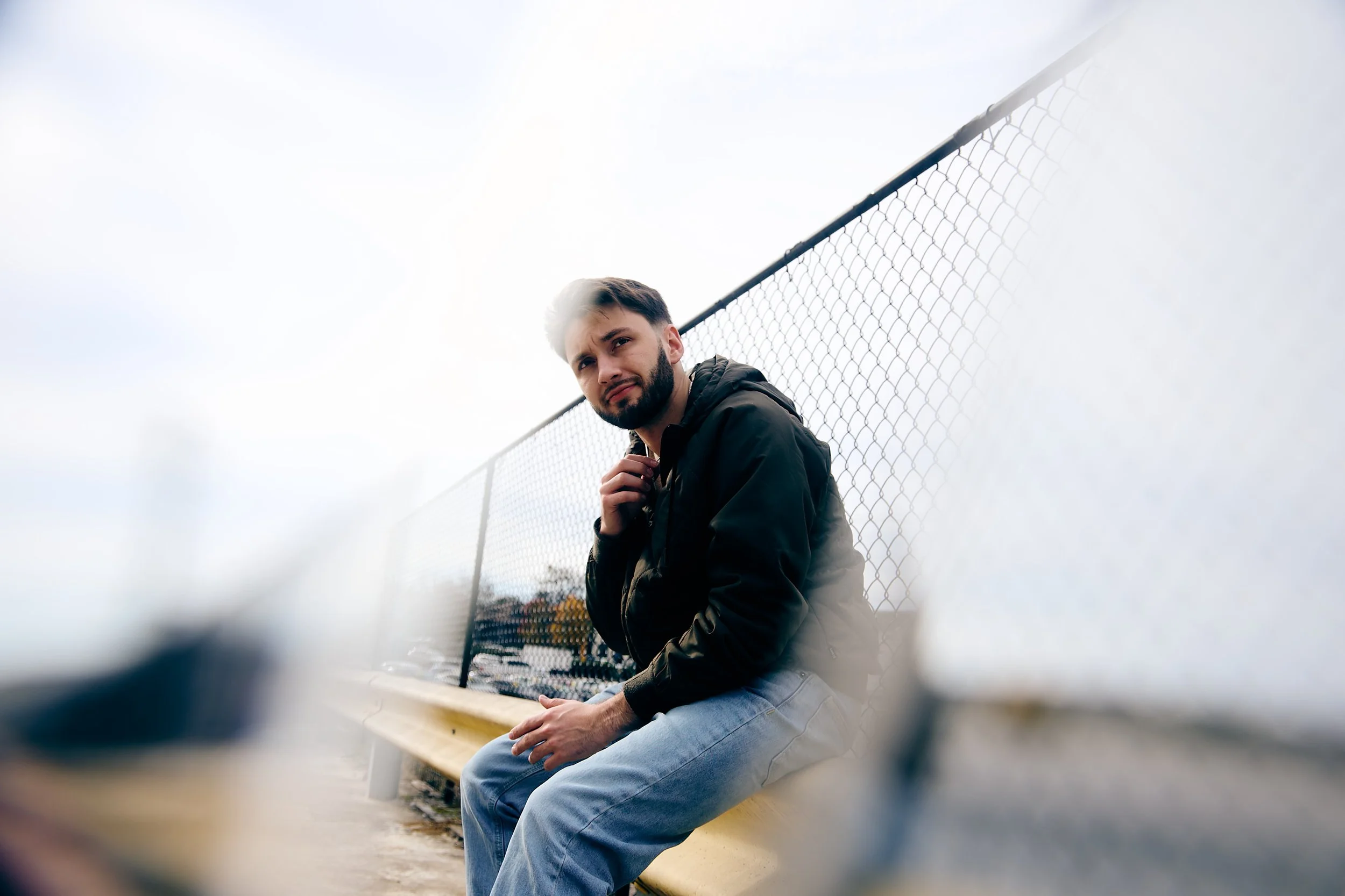 A man sitting on a yellow bench near a chain-link fence, wearing a black jacket and jeans, with a cloudy sky in the background.