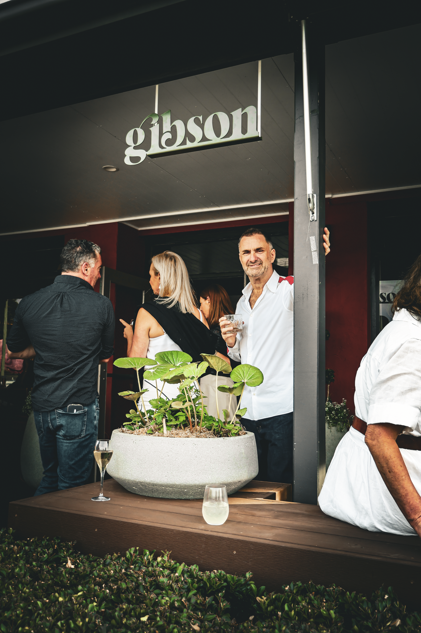People socializing outdoors at a restaurant under a sign that reads 'Gibson,' with a table holding a large pot with green plants, a glass of white wine, and a glass of clear liquid.
