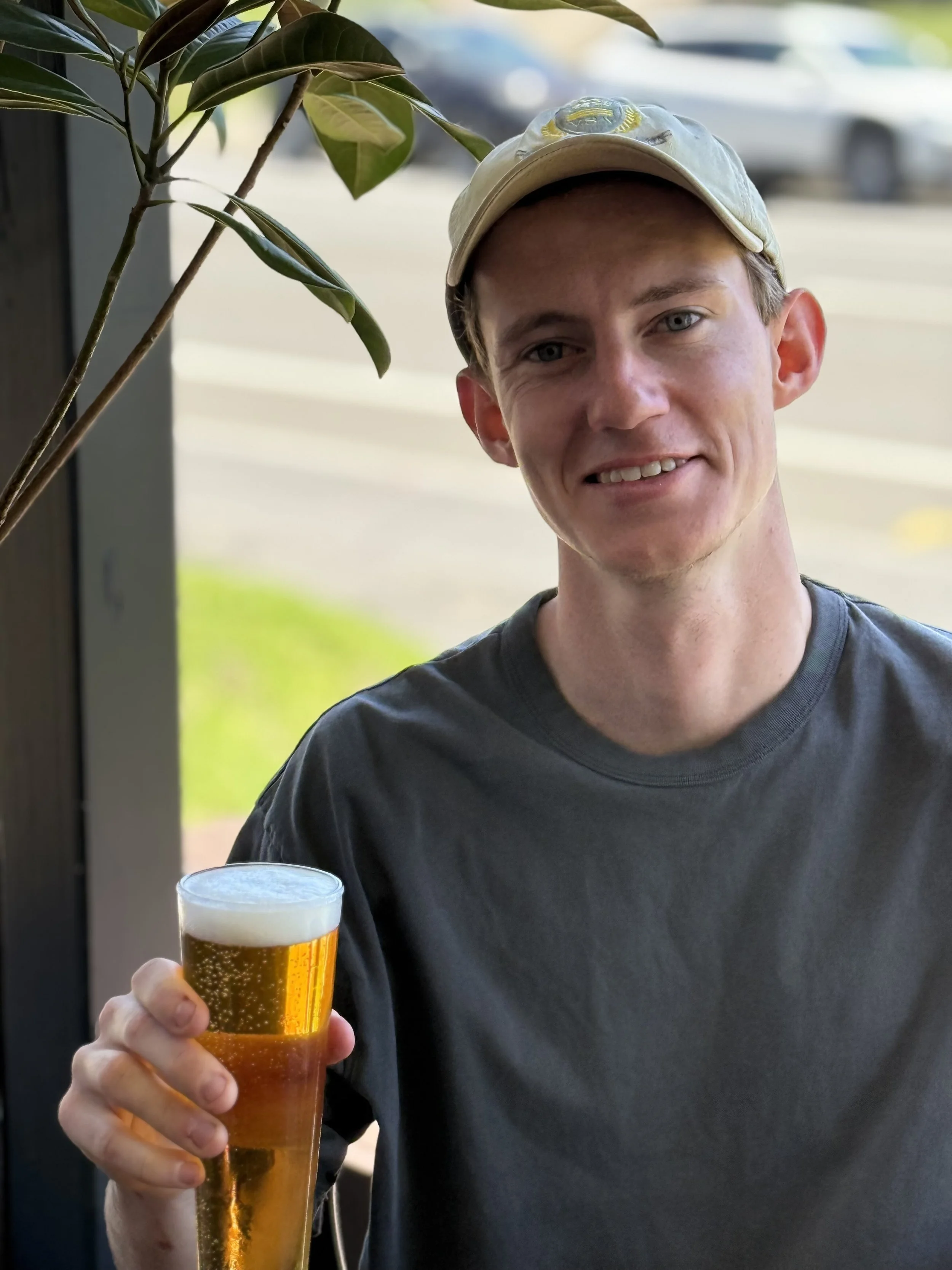 A young man with fair skin, blonde hair, wearing a baseball cap and a dark t-shirt, smiling while holding a glass of beer outside near some green plants and a blurred background of a parked car.