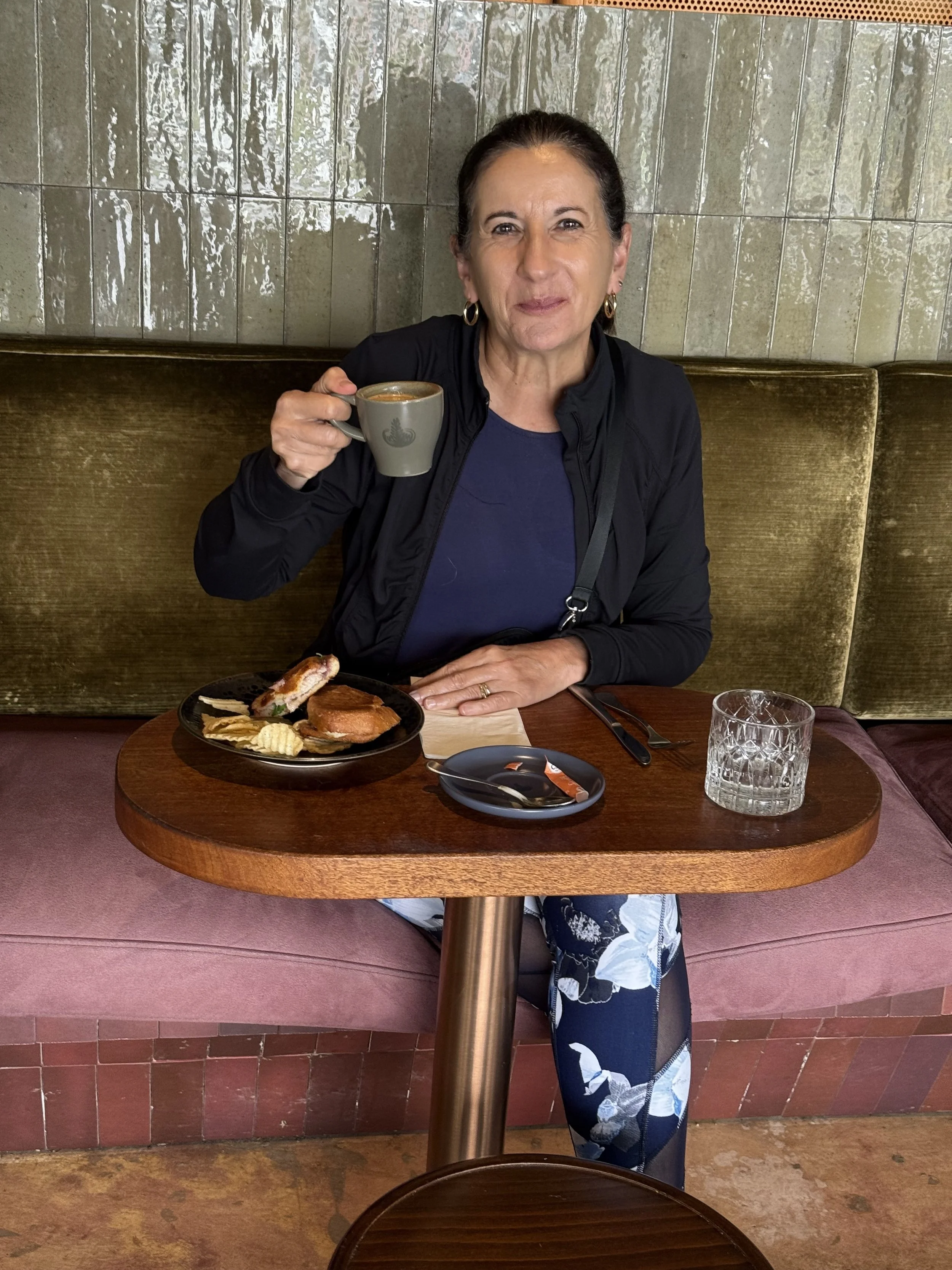 A woman sitting at a restaurant table holding a cup of coffee, with breakfast food on a plate and a glass of water in front of her.