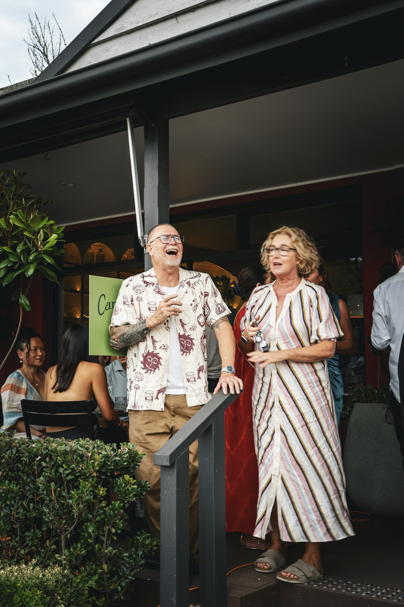 People gathered outdoors at a restaurant or bar, some standing and some sitting, laughing and talking. The background shows a building with large windows and a sign that says "Canti." The scene captures a lively social moment.