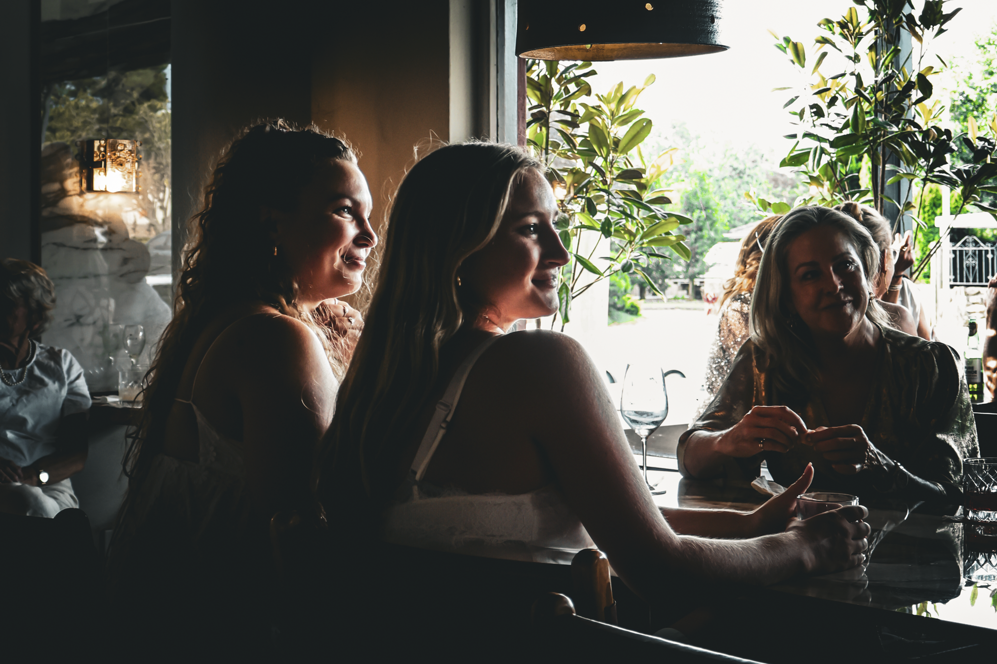 Four women sitting at a dining table inside a restaurant near a large window with natural light and greenery outside. They are smiling and engaging in conversation.