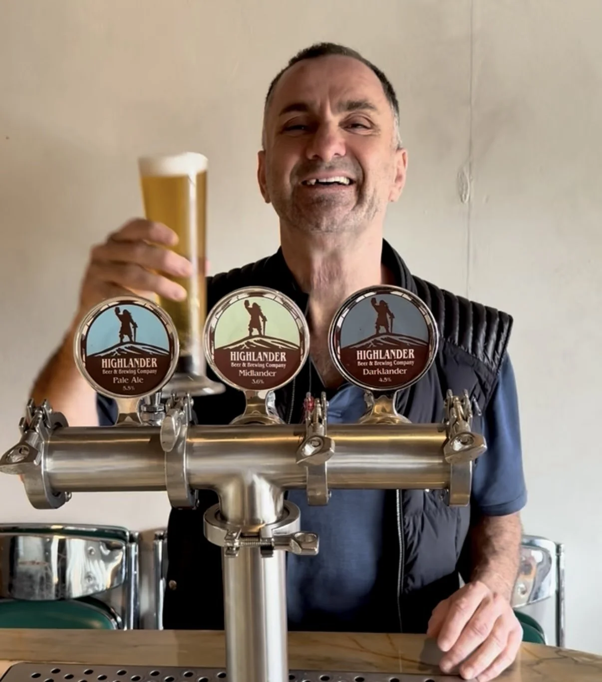 A man holding a glass of beer at a bar with Highlander beer tap handles in front of him.