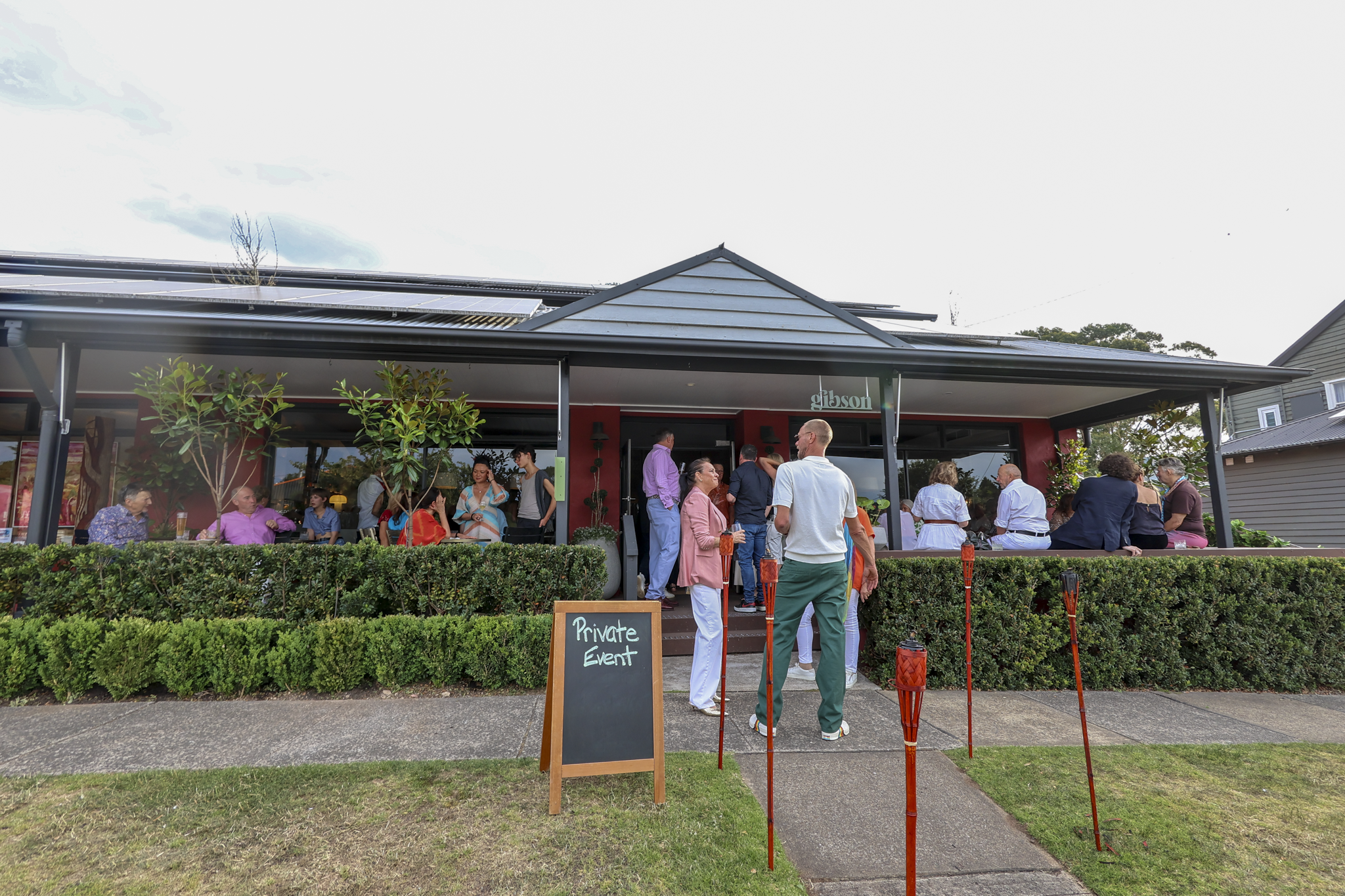 People gathered outside a restaurant with a sign that reads 'Private Event.' The restaurant has a large windows and is surrounded by greenery. Some individuals are standing and socializing, while others are seated inside visible through the windows.
