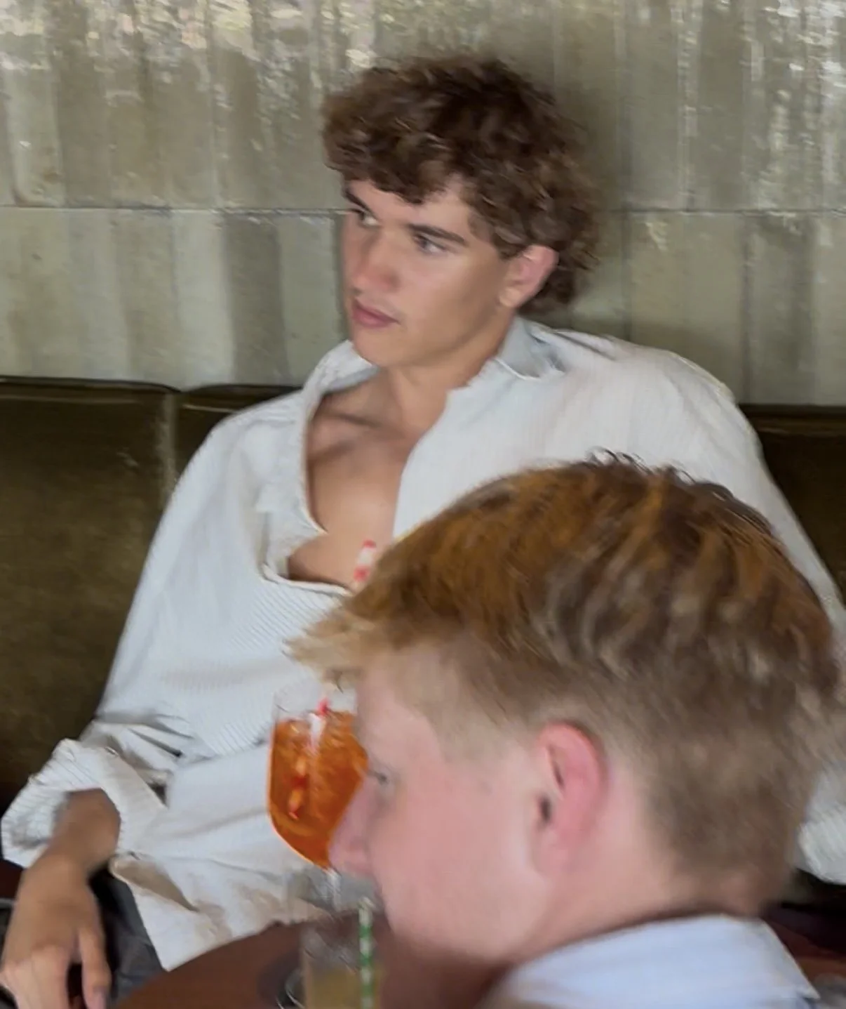 Two young men with reddish hair sitting at a table, one of them is drinking a beverage with a straw, and the other is looking to the side.