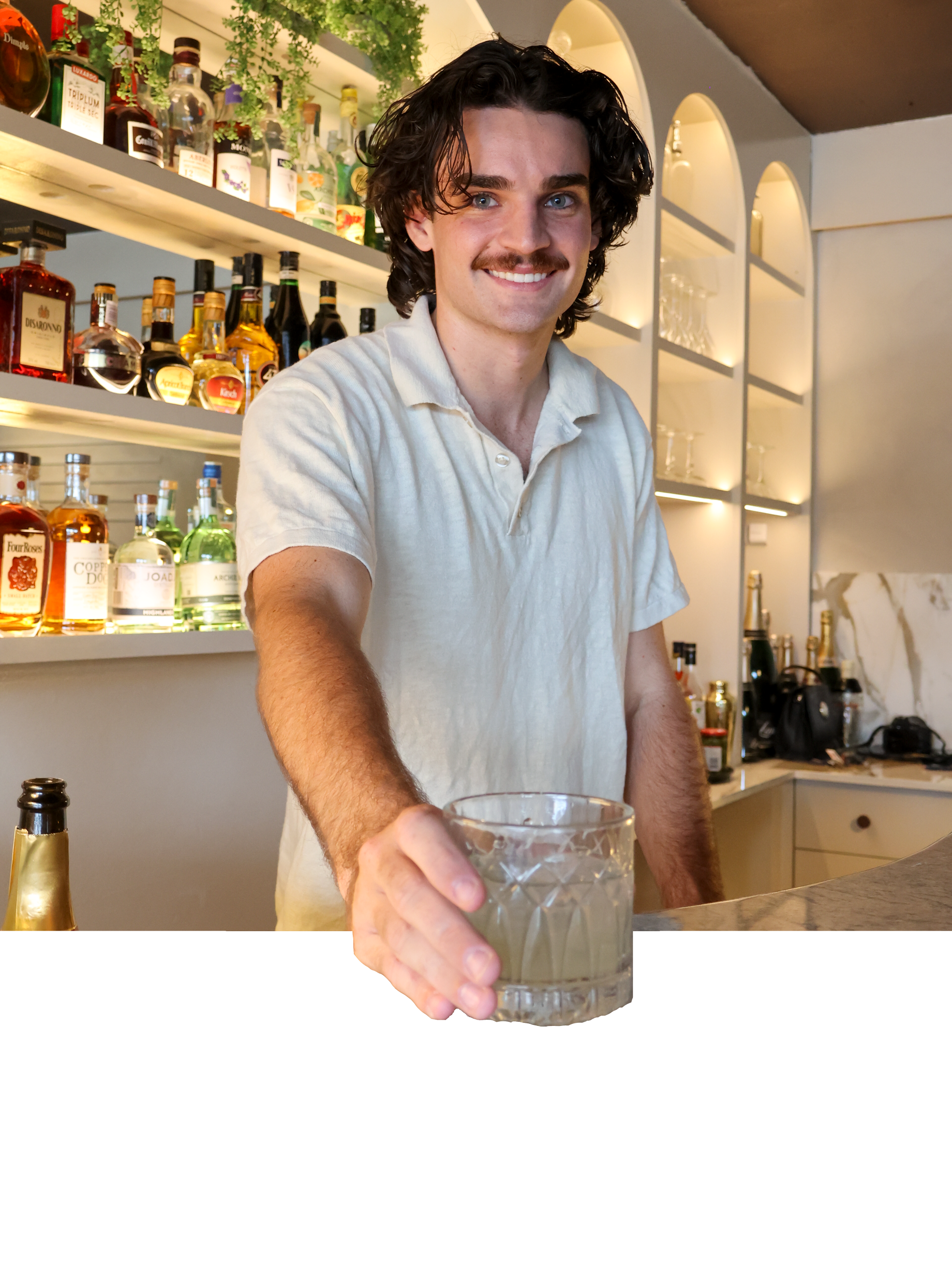 A man with dark curly hair, blue eyes, and a mustache holding a glass of drink is in a bar. The bar has shelves of alcohol bottles and barware, with warm lighting creating a cozy atmosphere.