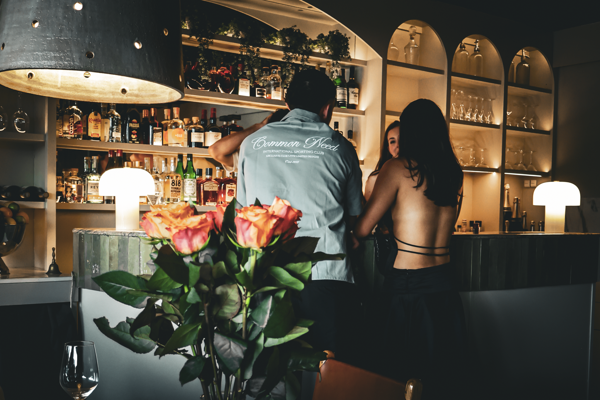 A dimly lit bar scene with a large flower arrangement in the foreground, a man and woman engaging in conversation at the bar, bottles of alcohol on shelves behind them, and a large lamp overhead.