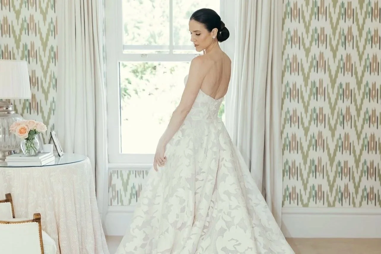 A woman in a white wedding dress standing in a bright room with patterned wallpaper, a window behind her, and a side table with flowers and books.